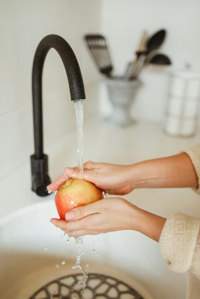 pexels-photo-5418582-5418582 Close-up of hands washing a red apple under a black kitchen faucet, symbolizing cleanliness.